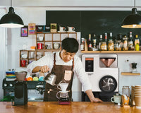 Person pouring coffee in a cafe with a Bellwether roaster behind them